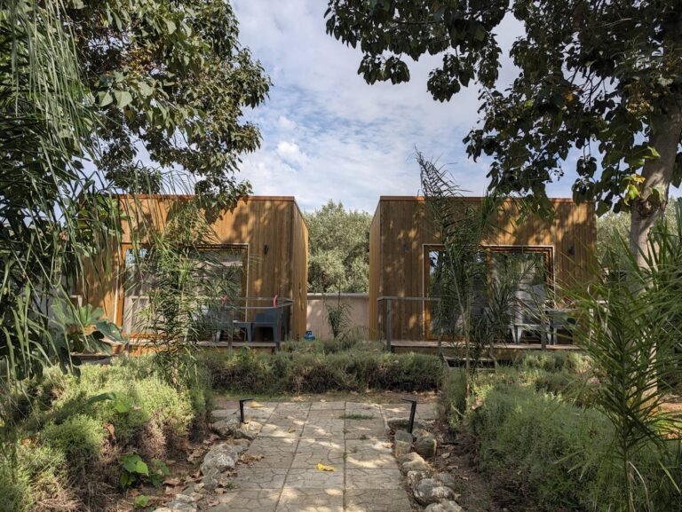 I due bungalow Cubo House visti dal vialetto del giardino, fra gli alberi e le piante